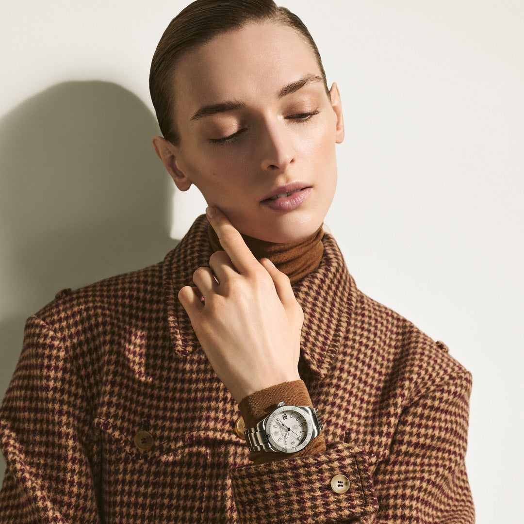 Woman wearing a brown houndstooth coat and a watch, posing against a plain background