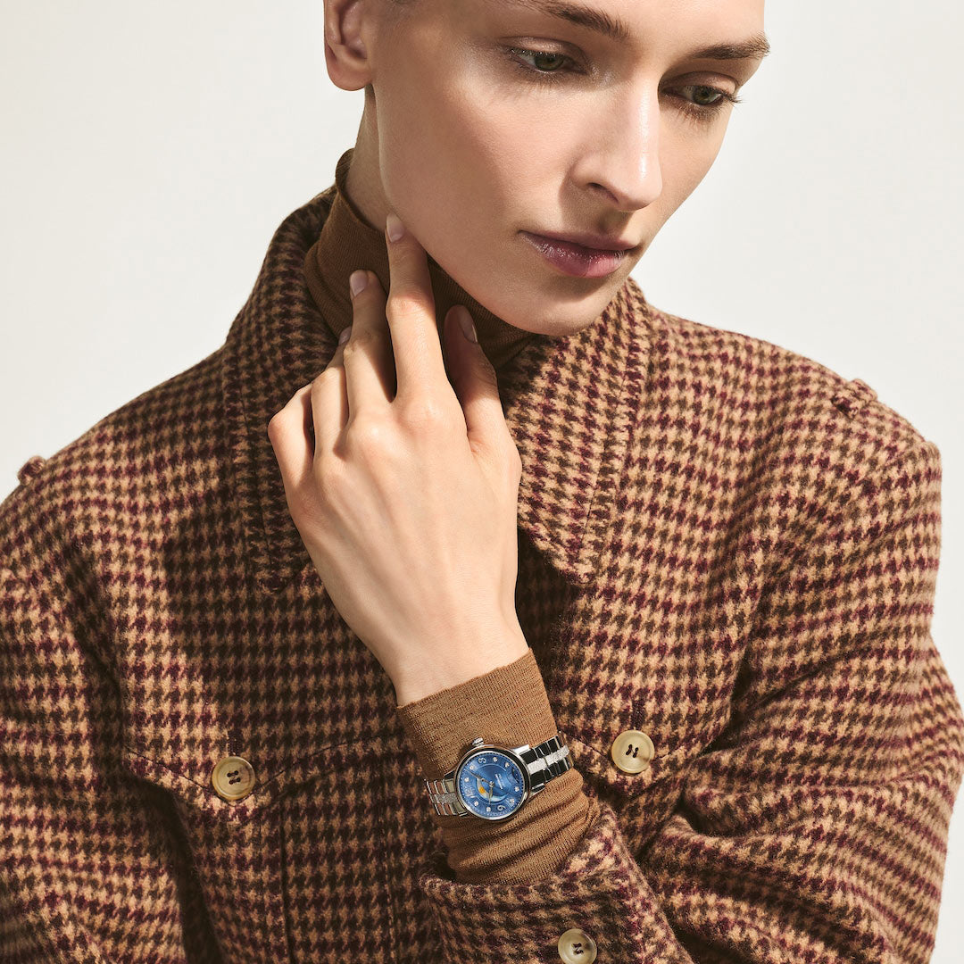 Person wearing a brown houndstooth coat with a watch on a plain background
