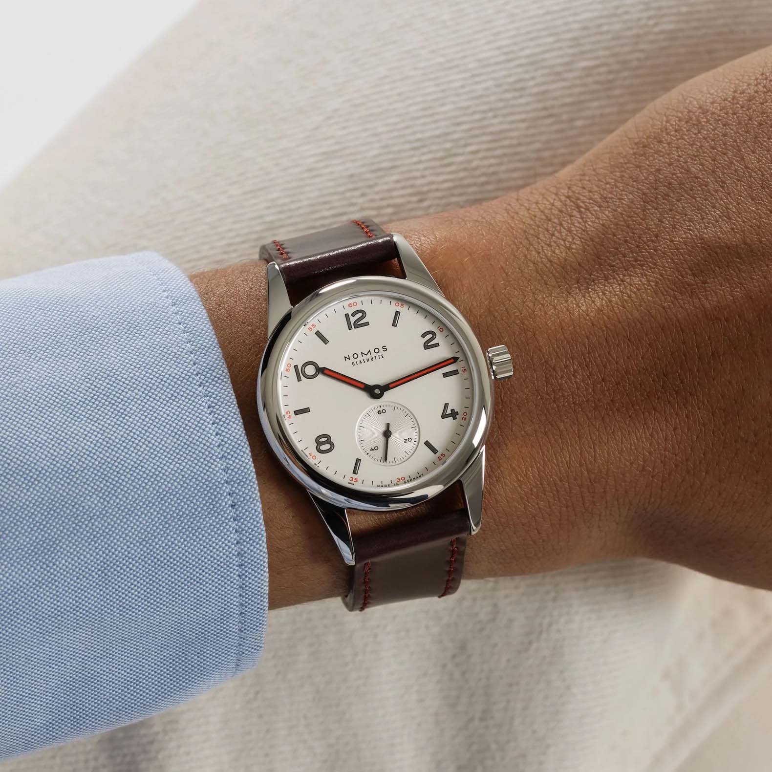 Man wearing NOMOS Glashütte Club 36mm watch with white silver-plated dial, black and red hands, and brown Horween leather strap