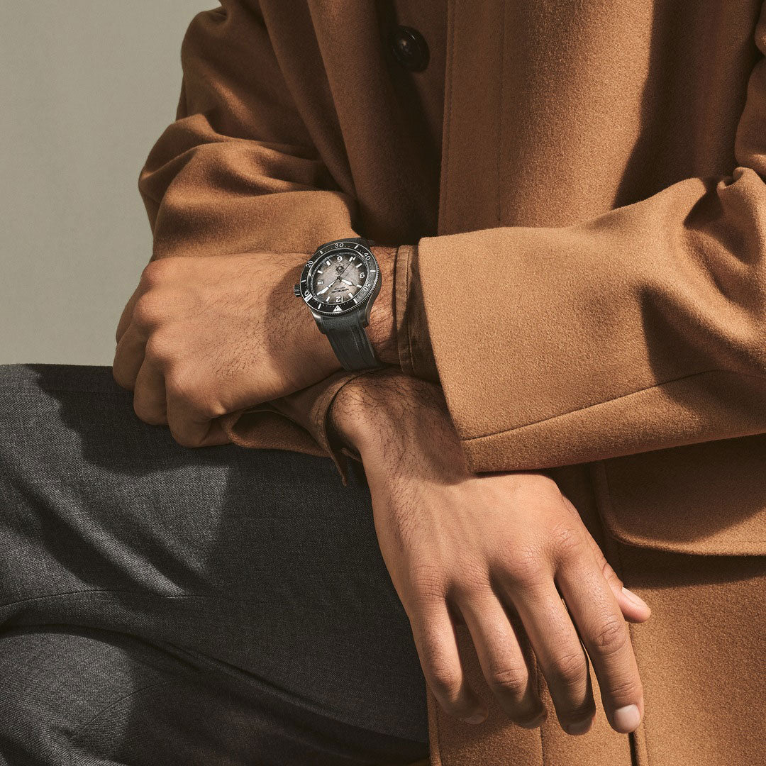 Person wearing a brown coat with a watch on their wrist, sitting against a neutral background