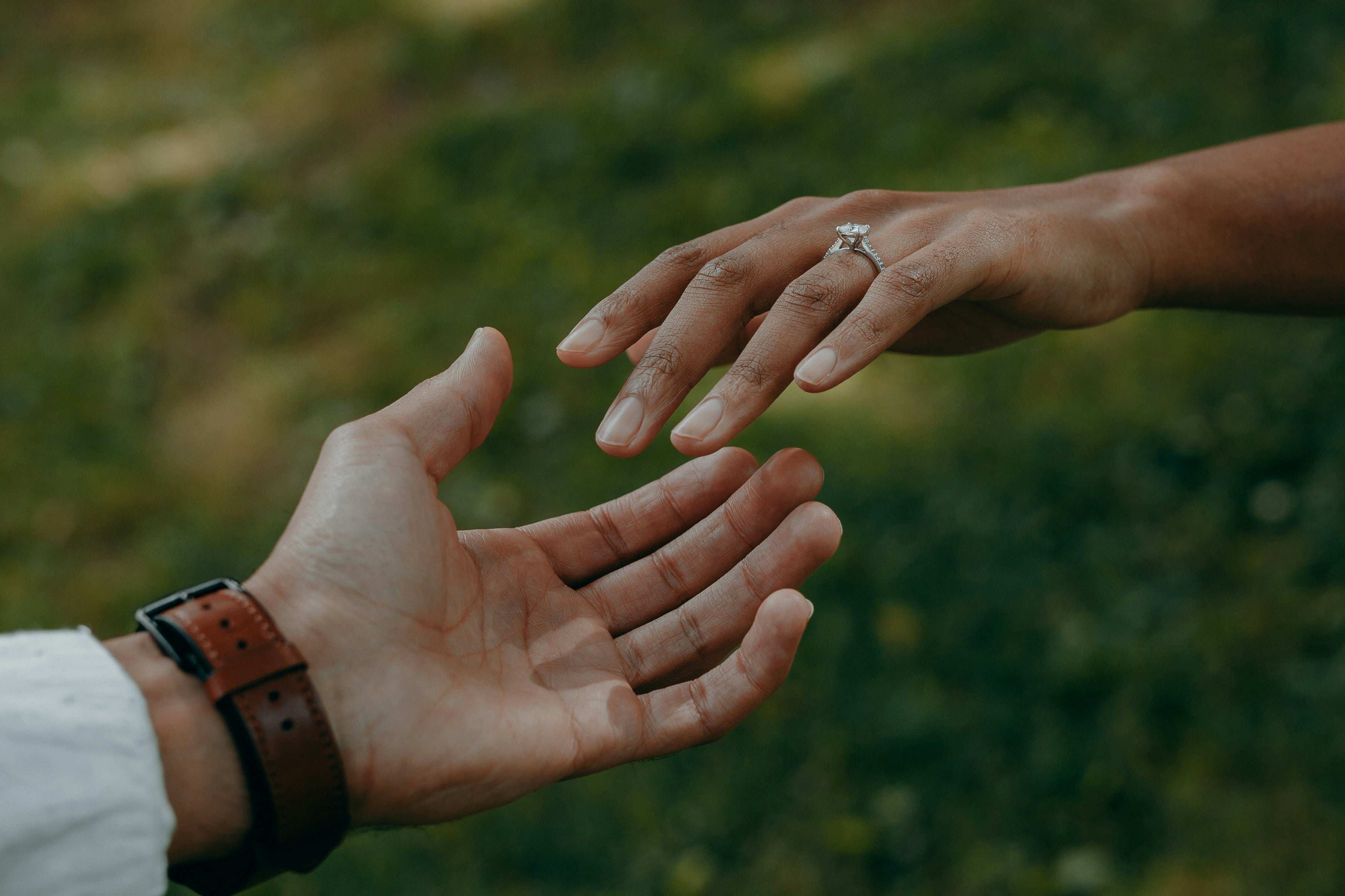 Two hands reaching towards each other with one hand wearing a ring, set against a blurred natural background.