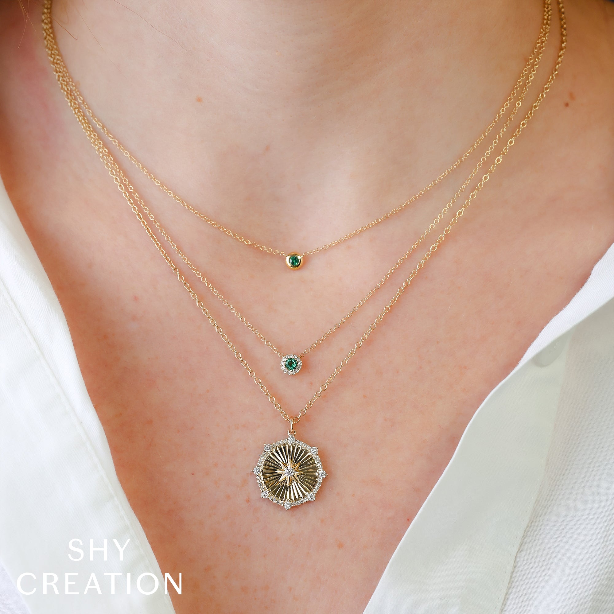 Gold necklaces with green emeralds on a person wearing a white top, branded 'SHY CREATION'.