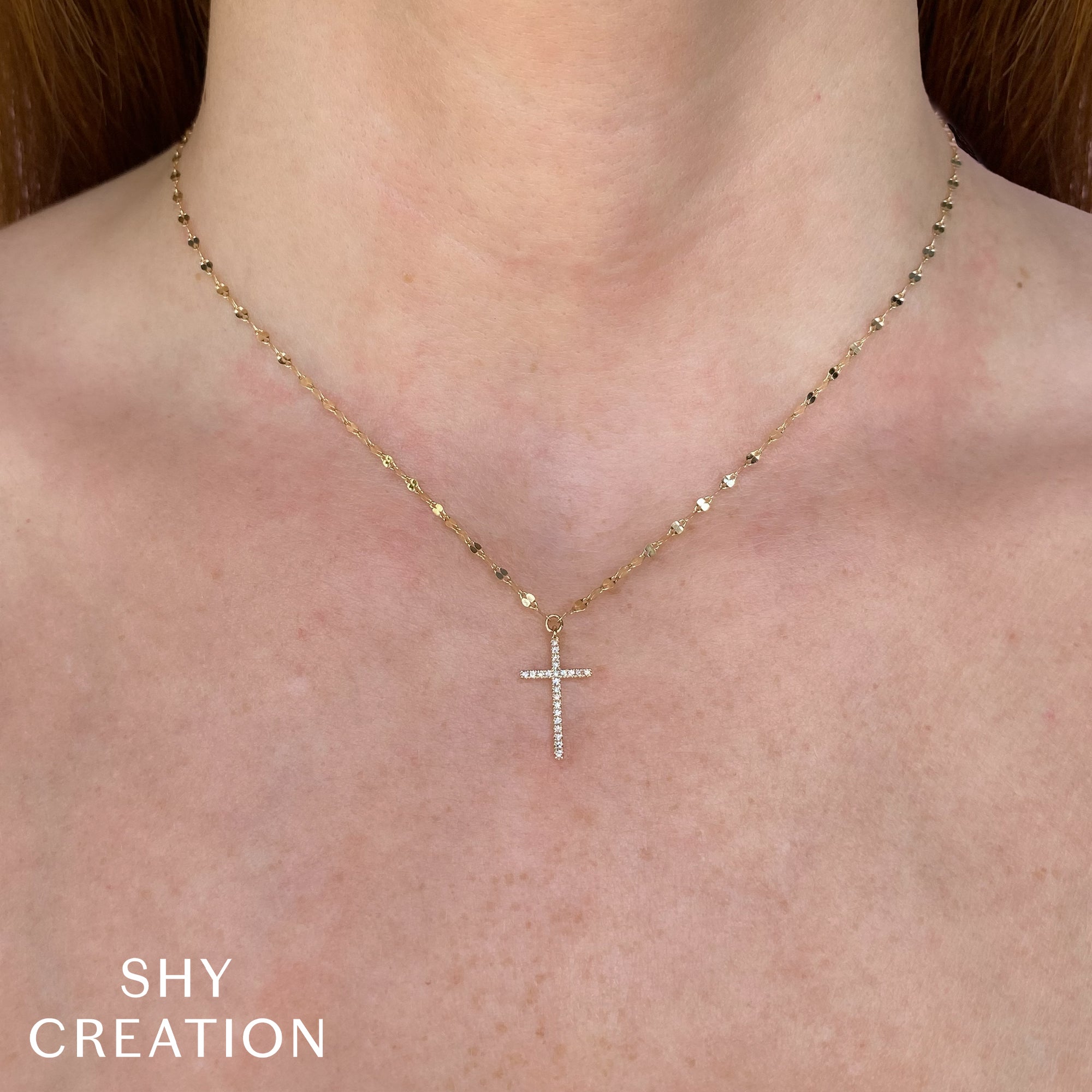 Gold necklace with a diamond cross pendant on a person's neck, branded 'SHY CREATION'.